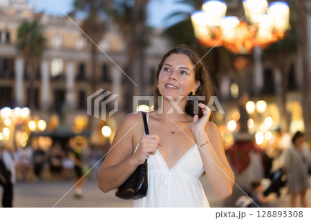 In evening, young smiling girl walks along city street Placa Real 128893308