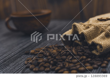 Burlap sack spilling coffee beans with blurred coffee cup background 128893369