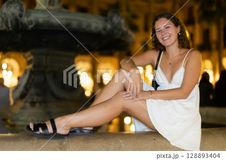 young woman sitting near a fountain in the evening 128893404
