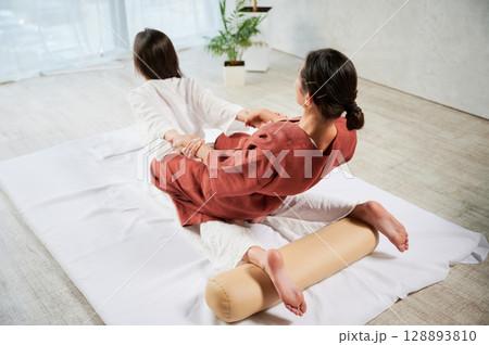 Masseuse making thai yoga massage. Woman therapist doing traditional massage treatment, stretching female patient's body. Client lying on mat, enjoying hand massage in calm atmosphere. 128893810