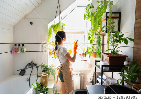 Portrait Woman in rubber gloves and apron manually cleaning in bathroom with rag cleaner and mop inside interior with home plants. Restoring order and cleanliness, cleaning servise 128893905