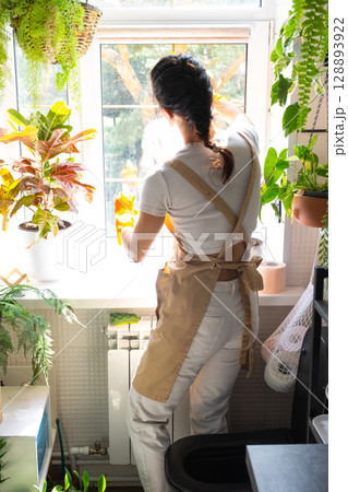 Woman in rubber gloves and apron manually washes window of house in bathroom with rag cleaner and mop inside interior with home plants. Restoring order and cleanliness, cleaning servise 128893922