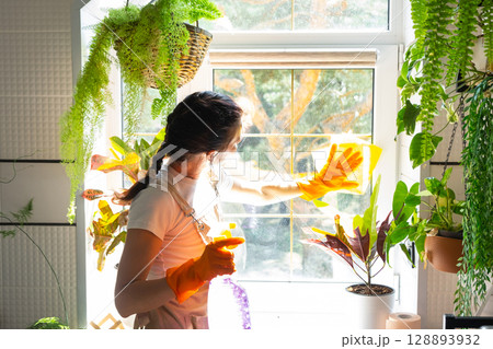 Woman in rubber gloves and apron close-up manually washes window of house with rag cleaner and mop inside interior with home plants on windowsill. Restoring order and cleanliness, cleaning servise Woman in rubber gloves and apron close-up manually washes window of house with rag cleaner and mop inside interior with home plants on windowsill. Restoring order and cleanliness, cleaning servise 128893932
