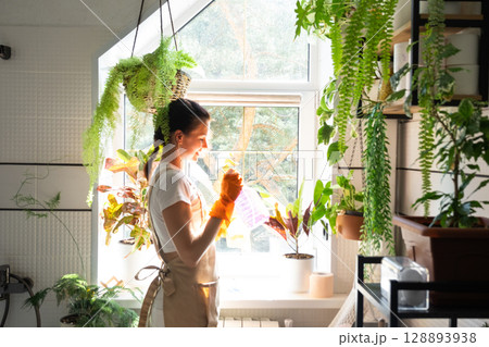 Woman in rubber gloves and apron manually washes window of house in bathroom with rag cleaner and mop inside interior with home plants. Restoring order and cleanliness, cleaning servise Woman in rubber gloves and apron manually washes window of house in bathroom with rag cleaner and mop inside interior with home plants. Restoring order and cleanliness, cleaning servise 128893938