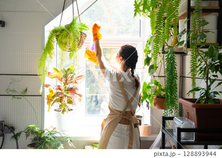 Woman in rubber gloves and apron manually washes window of house in bathroom with rag cleaner and mop inside interior with home plants. Restoring order and cleanliness, cleaning servise 128893944