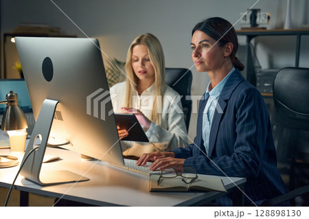 Professional Women Collaborating At Computer In Modern Office Setting At Night Professional Women Collaborating At Computer In Modern Office Setting At Night 128898130