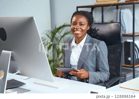 Confident Professional Woman Sitting at Desk Holding Tablet in Office Workspace Confident Professional Woman Sitting at Desk Holding Tablet in Office Workspace 128898164