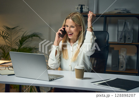 Excited Businesswoman Celebrating Success on Phone Call While Working at Office Desk Excited Businesswoman Celebrating Success on Phone Call While Working at Office Desk 128898207