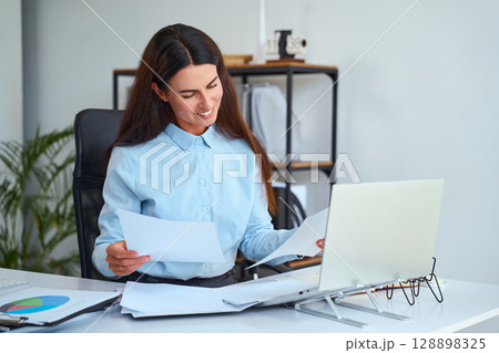 Smiling Businesswoman Reviewing Documents While Working on a Laptop in Modern Office 128898325