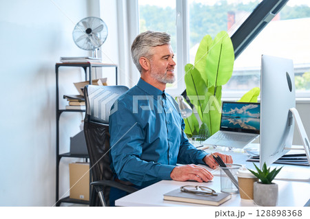 Professional Man Smiling While Working at Modern Office Desk With Computer Monitor 128898368