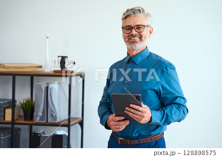 Smiling Professional Holding Tablet in Modern Office Space with Shelves in Background 128898575