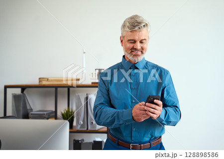 Smiling Mature Businessman Checking His Smartphone in a Modern Office Environment 128898586