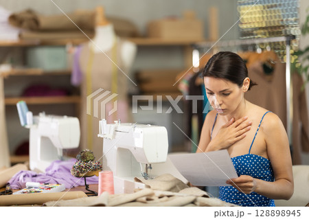 Upset young woman seamstress with documents in workshop Upset young woman seamstress with documents in workshop 128898945