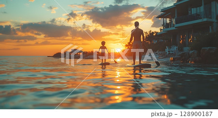 An adult and child paddleboard together on the ocean during a beautiful golden sunset near a tropical coastline. 128900187