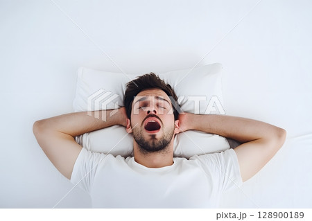 Young man yawns widely while lying comfortably in a clean white bed with hands behind his head. 128900189