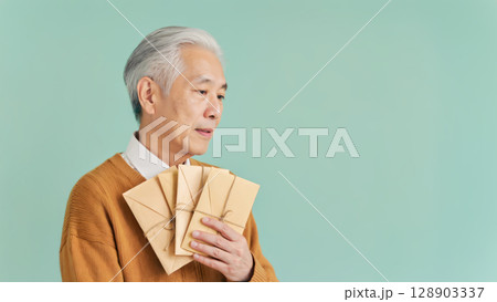 Asian elderly man holding traditional envelopes in warm-toned outfit against pastel background. Concept of cultural gifting, holiday traditions, generational care, and emotional connection visuals Asian elderly man holding traditional envelopes in warm-toned outfit against pastel background. Concept of cultural gifting, holiday traditions, generational care, and emotional connection visuals 128903337