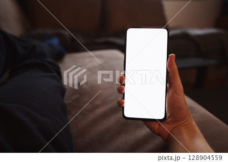 Man hold smartphone with white screen in his hand lying on a couch in home environment 128904559