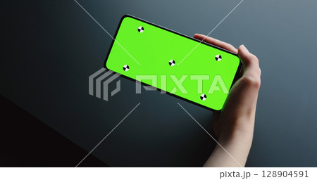 young woman sitting at the black table near window and holding smartphone with horizontal green screen 128904591