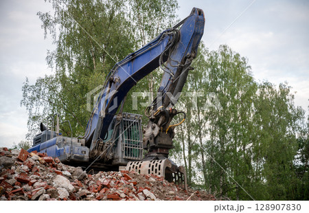 Blue excavator working on demolition site with pile of broken bricks and concrete rubble Blue excavator working on demolition site with pile of broken bricks and concrete rubble 128907830