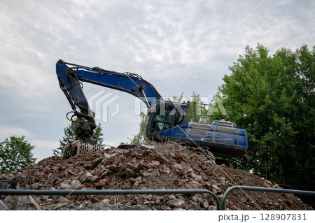 Blue excavator working on demolition site with pile of broken bricks and concrete rubble Blue excavator working on demolition site with pile of broken bricks and concrete rubble 128907831