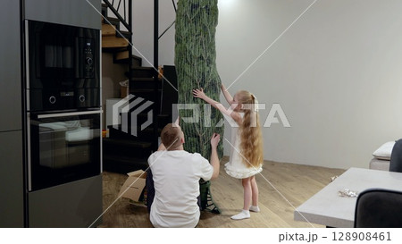 Father helping young daughter assembling artificial christmas tree, decorating living room with festive holiday spirit, bonding during seasonal family moment 128908461