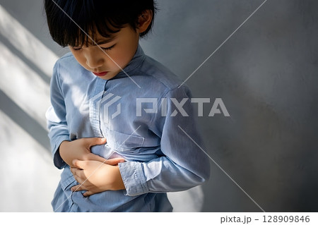 A young child experiences a stomach ache, standing alone indoors and clutching his abdomen with a look of discomfort. 128909846