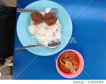 Plate of white rice with two meatballs and spoon, accompanied by bowl of spicy soup with meat and vegetables, served on blue table, showing simple meal with vibrant setting 128910384