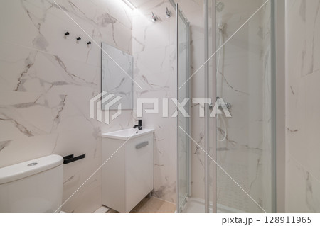 Modern bathroom with marble walls, glass shower, white sink, and toilet. Bright lighting enhances the clean, minimalist design. 128911965