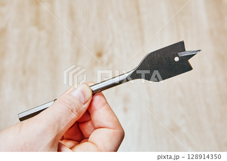 Flat drill bit with sharp cutting edges and small round hole near tip held on neutral background closeup shows woodworking drilling tool. 128914350