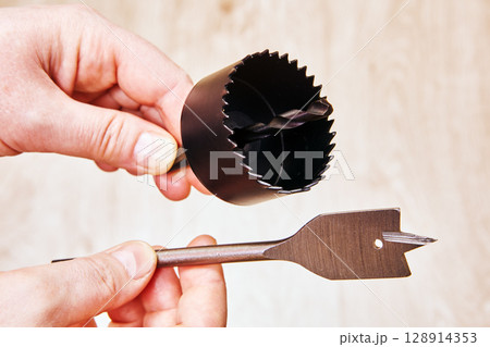 Spade bit and hole saw drill bit for woodworking held in hands close to each other ready to be attached to cordless drill for woodwork tasks. 128914353