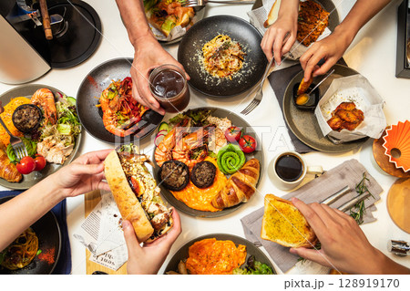 Captured from a bird's-eye view, five or six people are dining together at a Western-style restaurant, enjoying spaghetti, croissants, chicken wings, coffee, and other dishes. 128919170