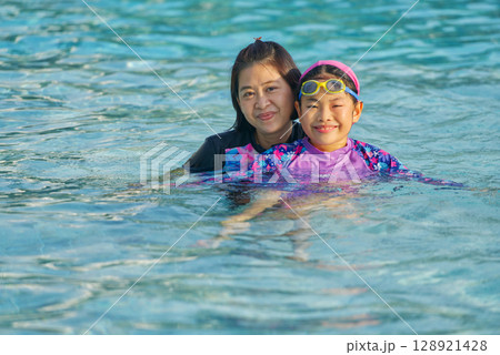 Portrait of a healthy middle-aged Asian mother and little daughter in a swimming pool, eyes looking at the camera, warm sunlight, space for copy and design. 128921428