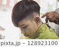 Barber Trimming Boy's Hair with Scissors and Comb in Barbershop 128923100
