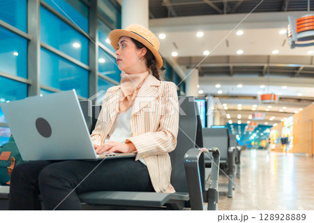 Bottom view of Caucasian elegant happy young woman is sitting on a chair in the waiting room of airport and working on a laptop. Concept of remote work 128928889