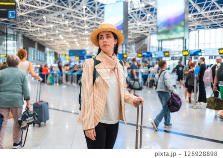 Pretty Caucasian young woman in straw hat wheeling a suitcase to the check-in desk. Defocused people in background. The concept of vacation and flight trip Pretty Caucasian young woman in straw hat wheeling a suitcase to the check-in desk. Defocused people in background. The concept of vacation and flight trip 128928898