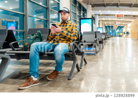 Wide shot of adult Caucasian man in cap and jacket using smartphone in airport waiting area sitting in chair. Low angle view. Concept of boarding at terminal before flight Wide shot of adult Caucasian man in cap and jacket using smartphone in airport waiting area sitting in chair. Low angle view. Concept of boarding at terminal before flight 128928899