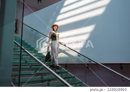 Wide view of a Caucasian beautiful young elegant woman in a straw hat and carrying a bag descending transparent glass steps. Sunbeams at the wall. Copy space 128928903