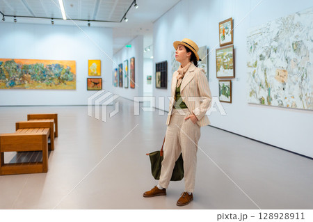 Museum day. Wide shot of young Caucasian pretty woman in elegant suit and straw hat posing in the art gallery. Concept of cultural education 128928911