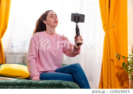 Mid shot of young woman recording an inspiring vlog for social media while sitting on a bed, using a smartphone on a tripod. 128928925