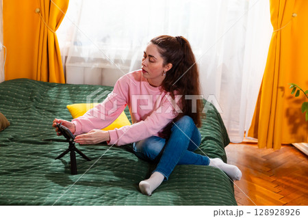 Mid shot of young Caucasian woman recording a social media vlog while sitting on a bed, discussing personal branding. Mid shot of young Caucasian woman recording a social media vlog while sitting on a bed, discussing personal branding. 128928926