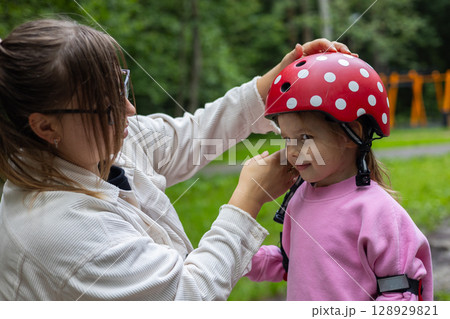 A young mother places a red helmet with white polka dots on her smiling daughter's head before riding a scooter in the park on a sunny summer day. Child safety while riding a scooter. 128929821
