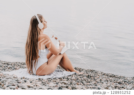 A woman is sitting on the beach with a laptop and headphones on. She is wearing a white bikini and she is enjoying her time by the water. A woman is sitting on the beach with a laptop and headphones on. She is wearing a white bikini and she is enjoying her time by the water. 128931252