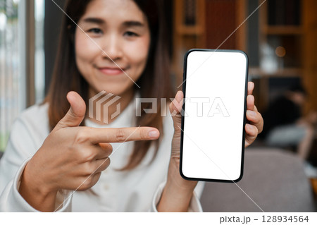 Smiling woman holding and pointing at a smartphone with a blank screen, perfect for mockup or advertisement use. Smiling woman holding and pointing at a smartphone with a blank screen, perfect for mockup or advertisement use. 128934564
