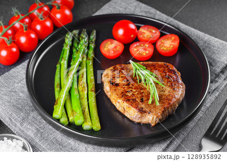 Grilled beef steak closeup with asparagus and cherry tomatoes on black plate, gourmet healthy dinner 128935892