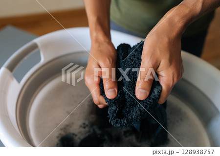 Close-up of handwashing dark clothing in plastic basin, wringing wet garment with bare hands over gray surface, showcasing manual laundry technique and water conservation at home. Close-up of handwashing dark clothing in plastic basin, wringing wet garment with bare hands over gray surface, showcasing manual laundry technique and water conservation at home. 128938757