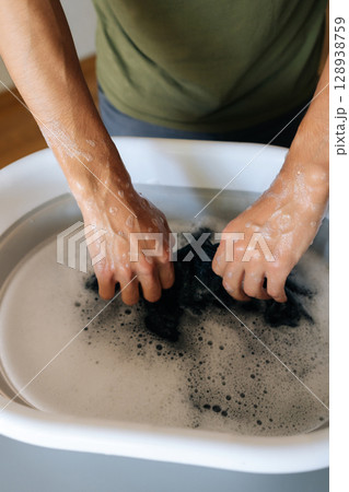 Vertical cropped shot of unrecognizable man soaking black laundry in water in folding basin standing at gray table doing household chores. Concept of hygiene and cleanliness in domestic environment. 128938759