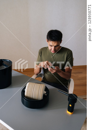 Vertical portrait of puzzled man cleaning air humidifier filter, referencing smartphone tutorial, showing disassembled device components on table surface. Concept of maintenance modern home appliance. 128938771