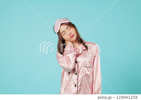 Asian woman experiences neck and shoulder pain, feeling tired needing sleep, wearing pajamas and sleep mask, possibly stretching after long sleep, headshots isolated studio blue background Asian woman experiences neck and shoulder pain, feeling tired needing sleep, wearing pajamas and sleep mask, possibly stretching after long sleep, headshots isolated studio blue background 128941258