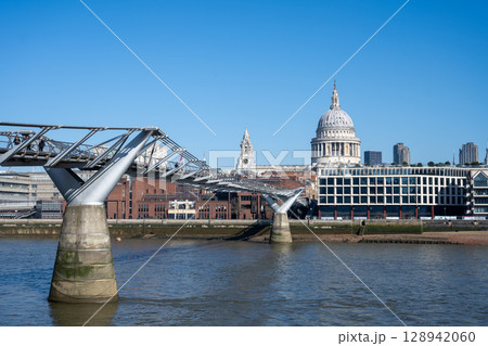 Millennium Bridge connects areas of London over the River Thames, with the iconic St. Paul's Cathedral visible in the background under a clear blue sky. People enjoy the view. 128942060