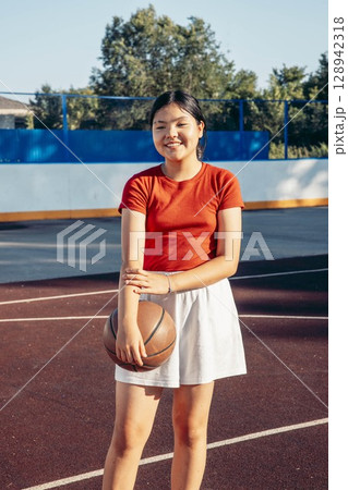 smiling Asian teenage girl in red shirt, white skirt holding basketball on outdoor court on sunny summer day, youth sport girl empowerment healthy lifestyle and leisure physical activity smiling Asian teenage girl in red shirt, white skirt holding basketball on outdoor court on sunny summer day, youth sport girl empowerment healthy lifestyle and leisure physical activity 128942318